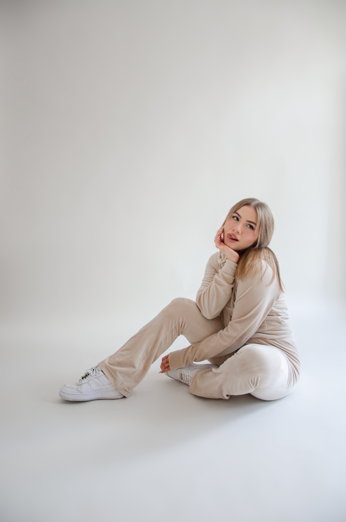 Młoda kobieta w beżowym welurowym dresie i białych sportowych butach, siedząca na podłodze na jasnym tle, z ręką podpierającą brodę i językiem wystawionym w bok.