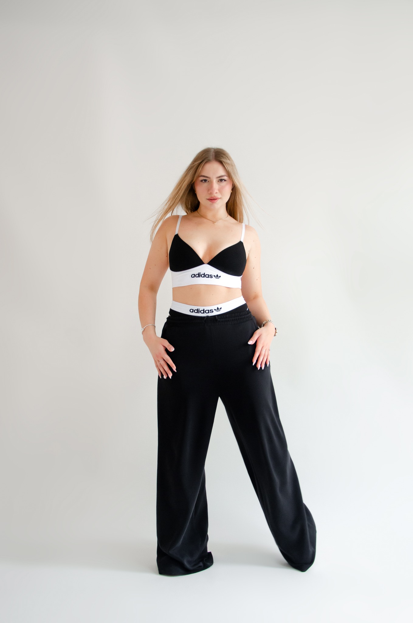 Portret kobiety w sportowym biustonoszu i szerokich spodniach dresowych, na białym tle, z delikatnym uśmiechem i naturalnym makijażem.