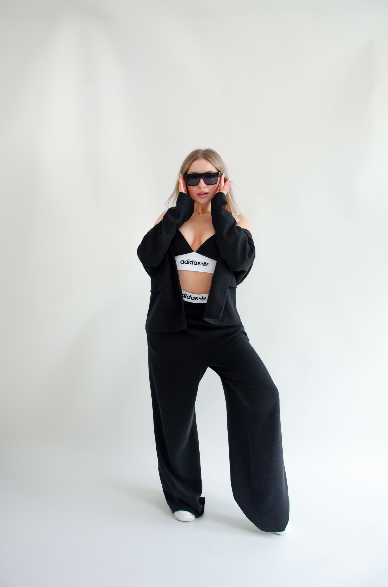 Kobieta w czarnym garniturze i okularach przeciwsłonecznych pozuje na białym tle, ubrana w sportowy top i spodnie z logo Adidas.