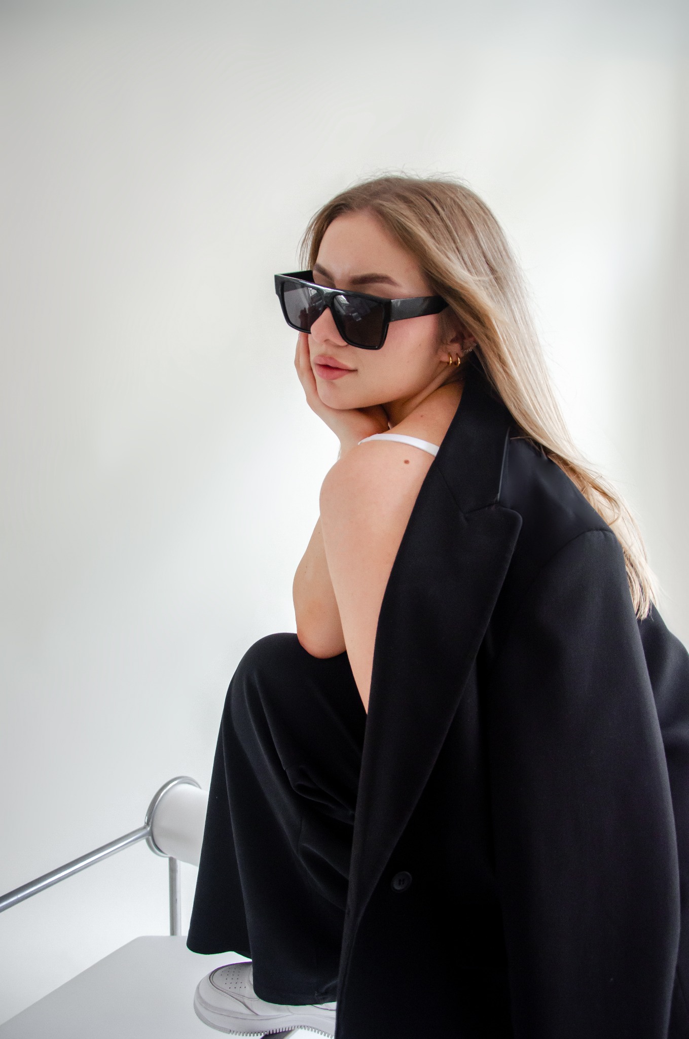 Portret kobiety w czarnym garniturze i okularach przeciwsłonecznych, opierającej brodę na dłoni, siedzącej na białym krześle na tle jasnej ściany.