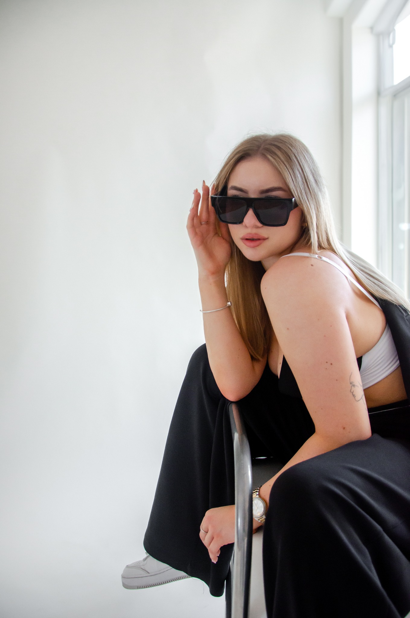 Portret kobiety w czarnym kombinezonie i okularach przeciwsłonecznych, siedzącej na krześle na tle białej ściany i okna, z widocznym tatuażem na ramieniu i zegarkiem na ręce.
