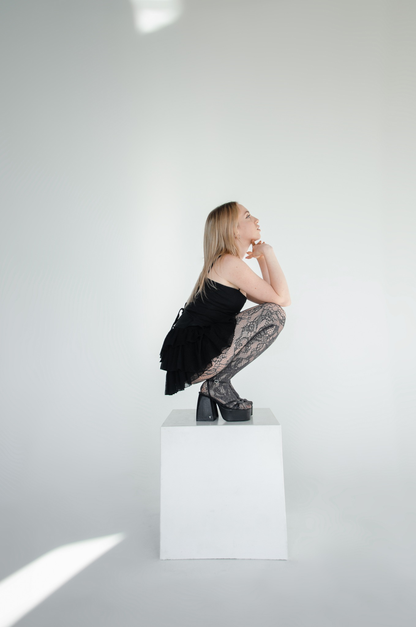Młoda blondynka w czarnej sukience i wzorzystych rajstopach, siedząca w kucki na białym sześcianie, z rękami pod brodą, patrzy w górę na białym tle.