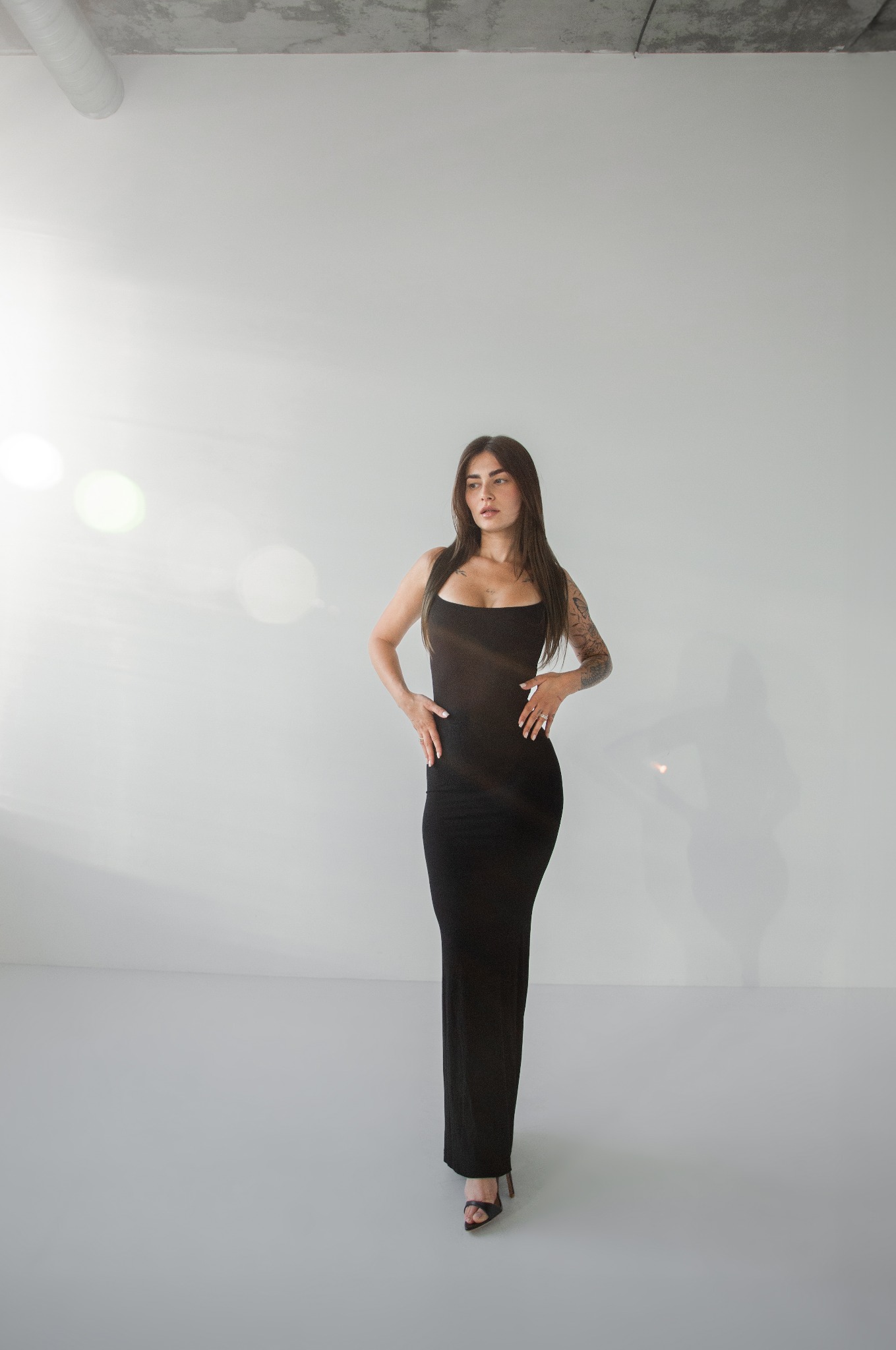 Kobieta w czarnej, dopasowanej sukience na ramiączkach, z tatuażem na ramieniu, pozuje w minimalistycznym, jasnym studio z betonowym sufitem.