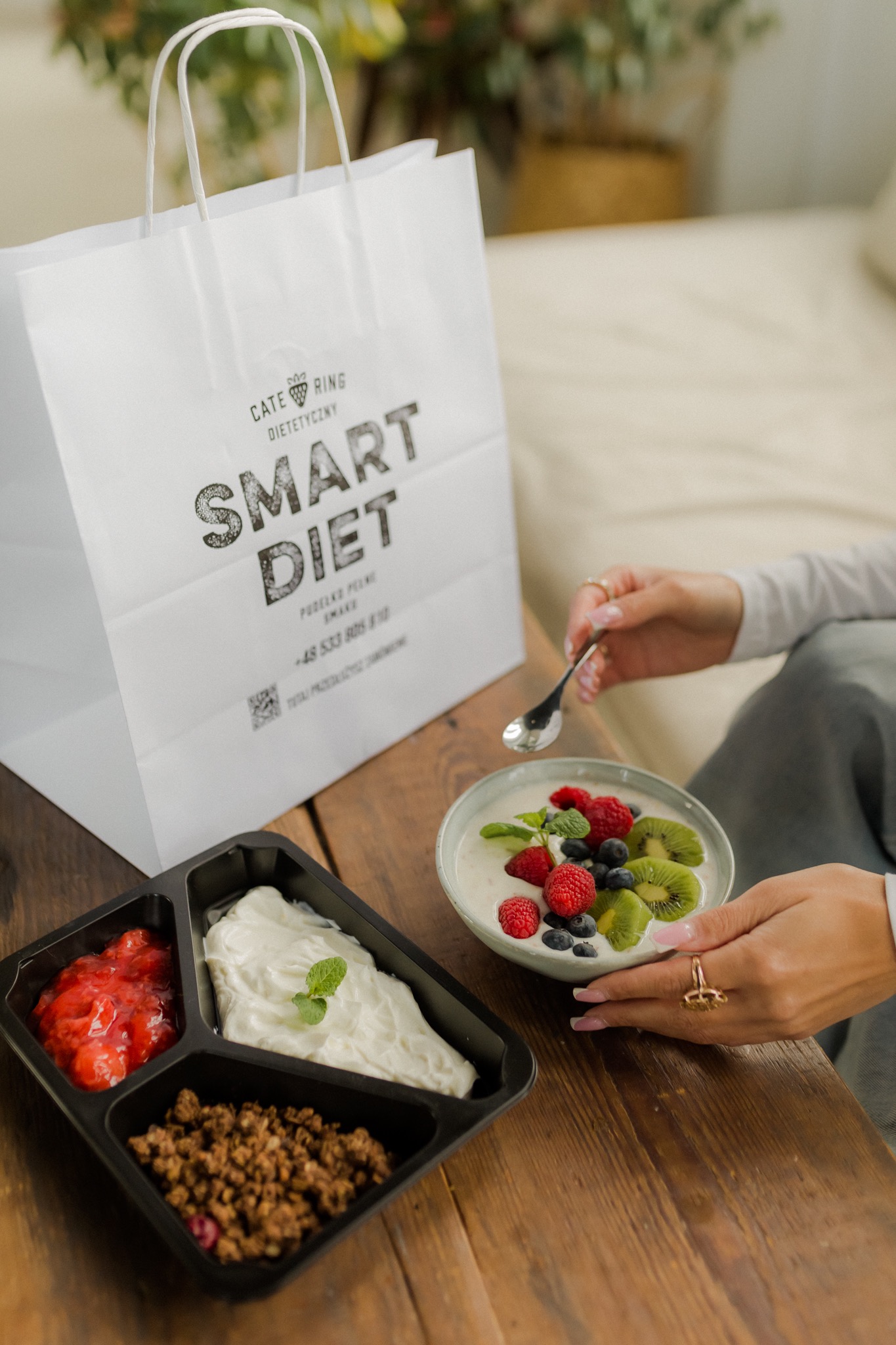 Osoba spożywa jogurt z owocami z miseczki, obok pudełko z trzema przegródkami zawierające dżem truskawkowy, jogurt naturalny i granolę, a w tle torba papierowa z logo firmy cateringowej.