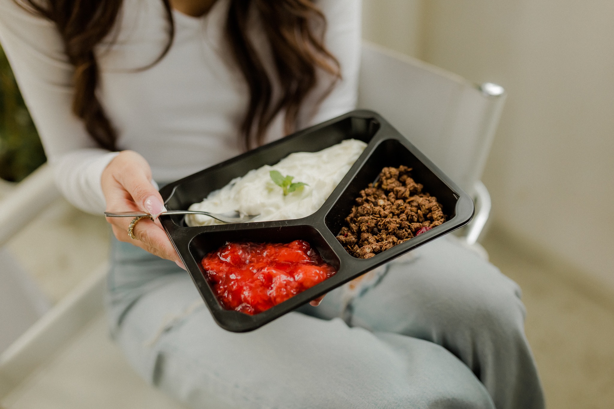 Kobieta trzyma czarne pudełko z trzema przegródkami zawierającymi: białą masę z miętą, brązowe granulki i czerwoną konfiturę, siedząc na krześle.