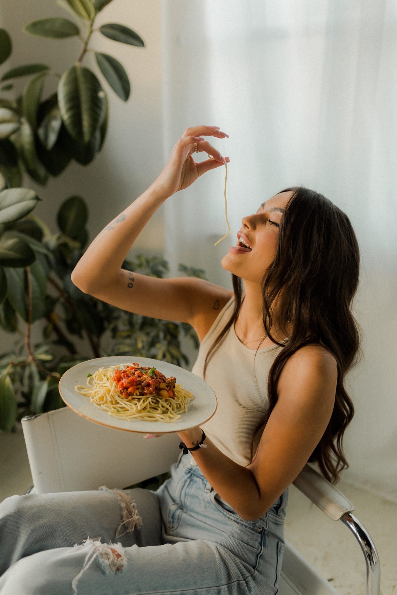 Uśmiechnięta kobieta siedzi z talerzem spaghetti z sosem pomidorowym, podnosząc pojedynczy makaron do ust, w tle duża roślina doniczkowa i minimalistyczne wnętrze.