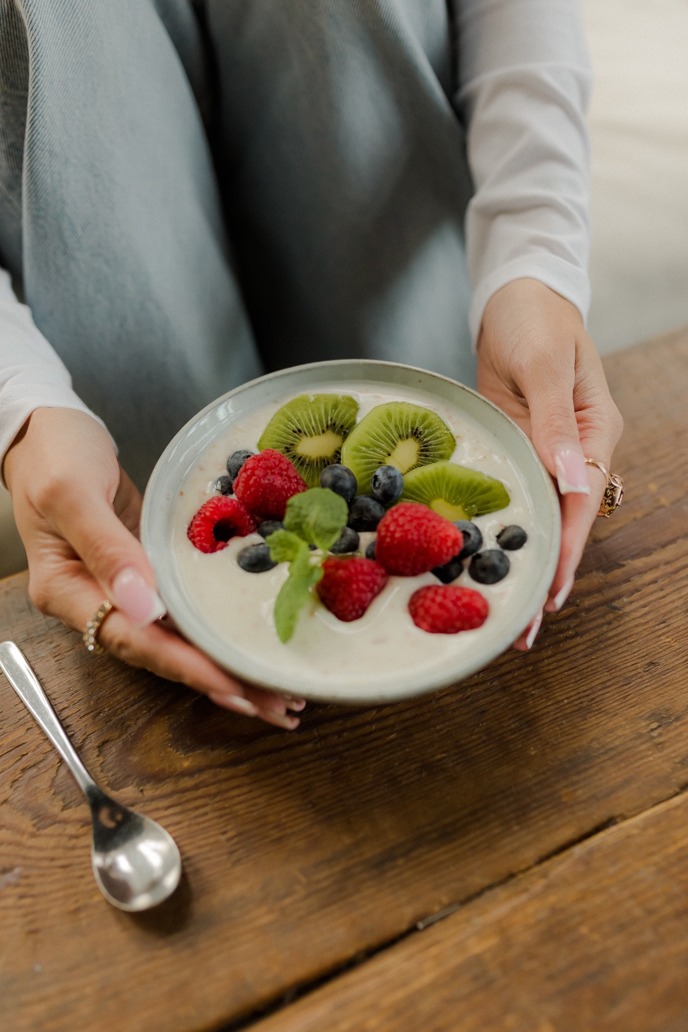 Owsianka z kiwi, malinami i borówkami, trzymana w dłoniach z pierścionkami, na drewnianym stole obok łyżeczki.