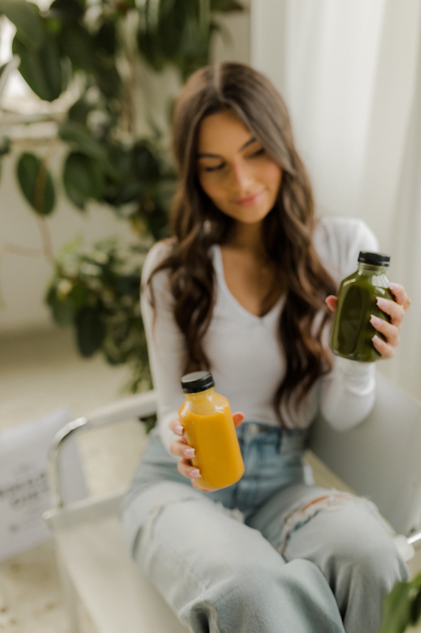 Kobieta siedzi na krześle, trzymając w dłoniach dwie butelki: jedną z pomarańczowym, a drugą z zielonym napojem. W tle widoczne rośliny doniczkowe i jasna zasłona.