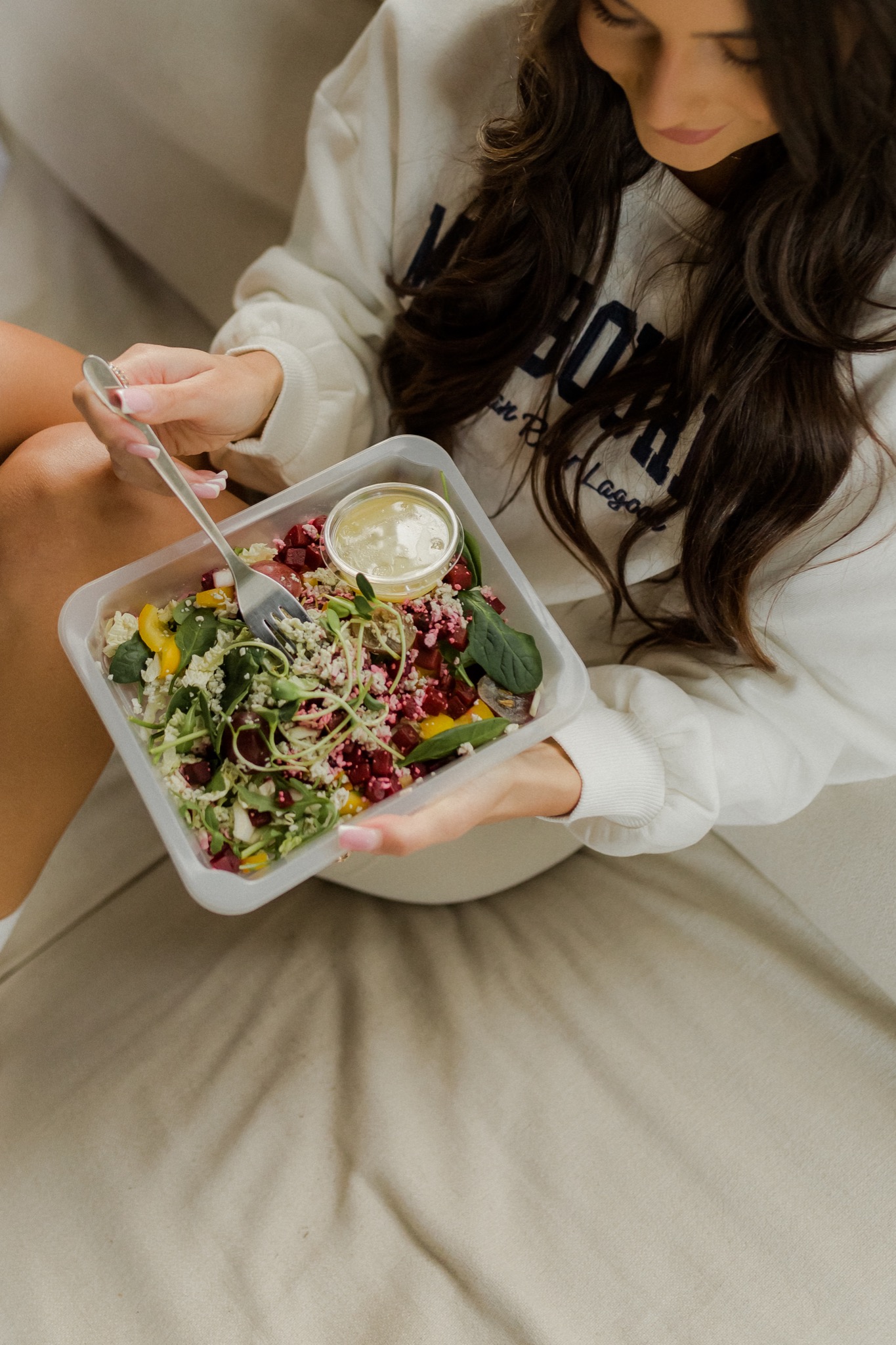 Kobieta w białej bluzie je sałatkę z burakiem, serem, sałatą i kiełkami z plastikowego pojemnika, trzymając widelec w dłoni z manicure.