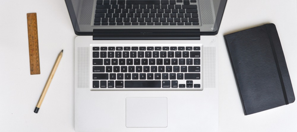 Widok z góry na otwarty laptop, notatnik, ołówek i drewnianą linijkę na białym biurku. Sprzęt biurowy w neutralnej tonacji. Minimalizm.