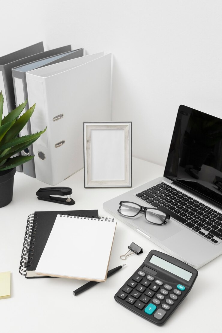 Biuro z laptopem, kalkulatorem, notatnikami, segregatorami i ramką na zdjęcia. Okulary leżą na laptopie. Minimalistyczne, białe biurko.