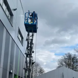 Pracownik na podnośniku koszowym myje elewację budynku biurowego pod ciśnieniem, widoczne okna i lampy na ścianie, pochmurne niebo w tle.