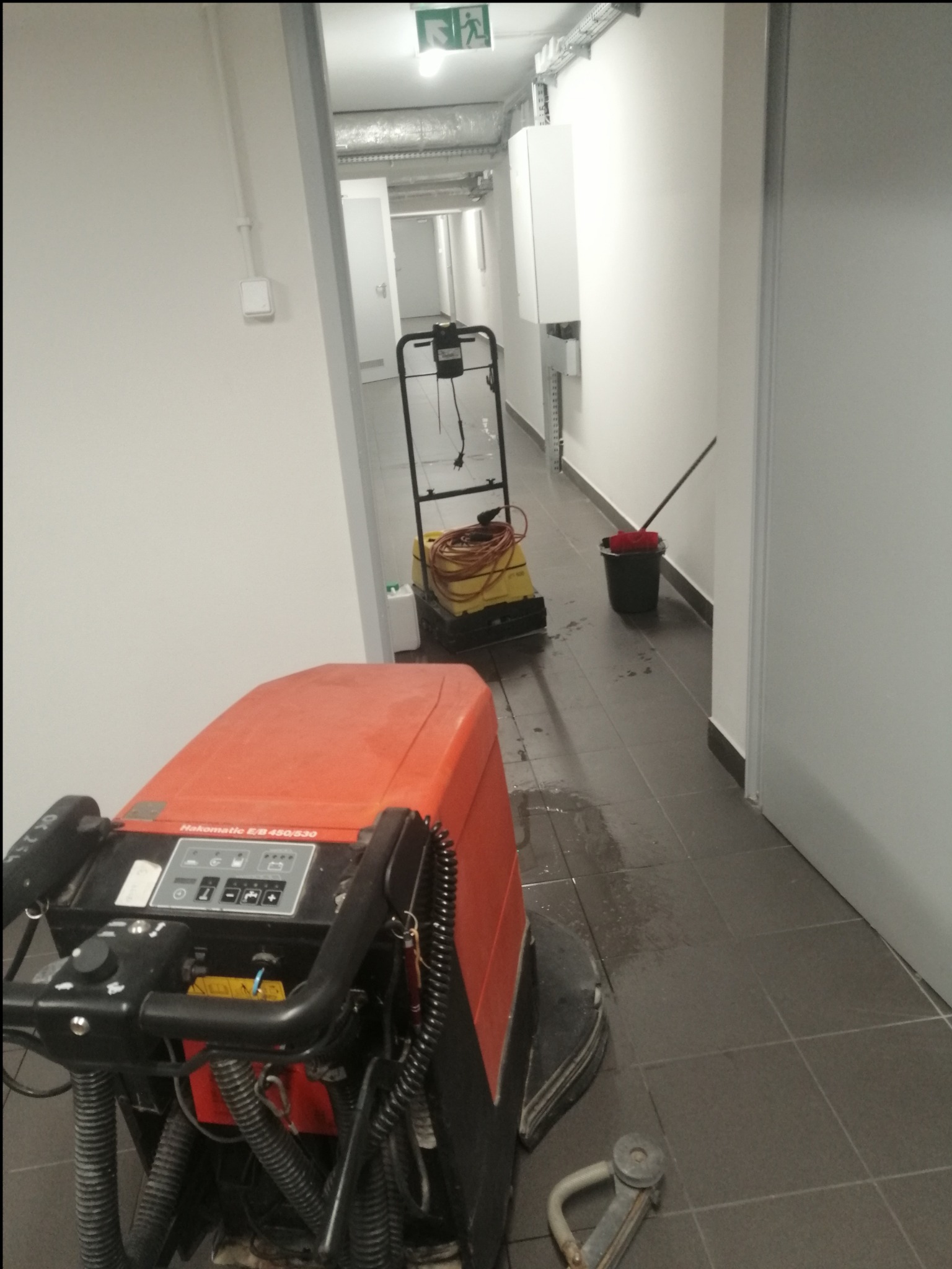Pomarańczowa maszyna czyszcząca Hakomatic EB 450/530 na mokrej, szarej posadzce w korytarzu biurowym, w tle wiadro z mopem i kolejna maszyna czyszcząca.