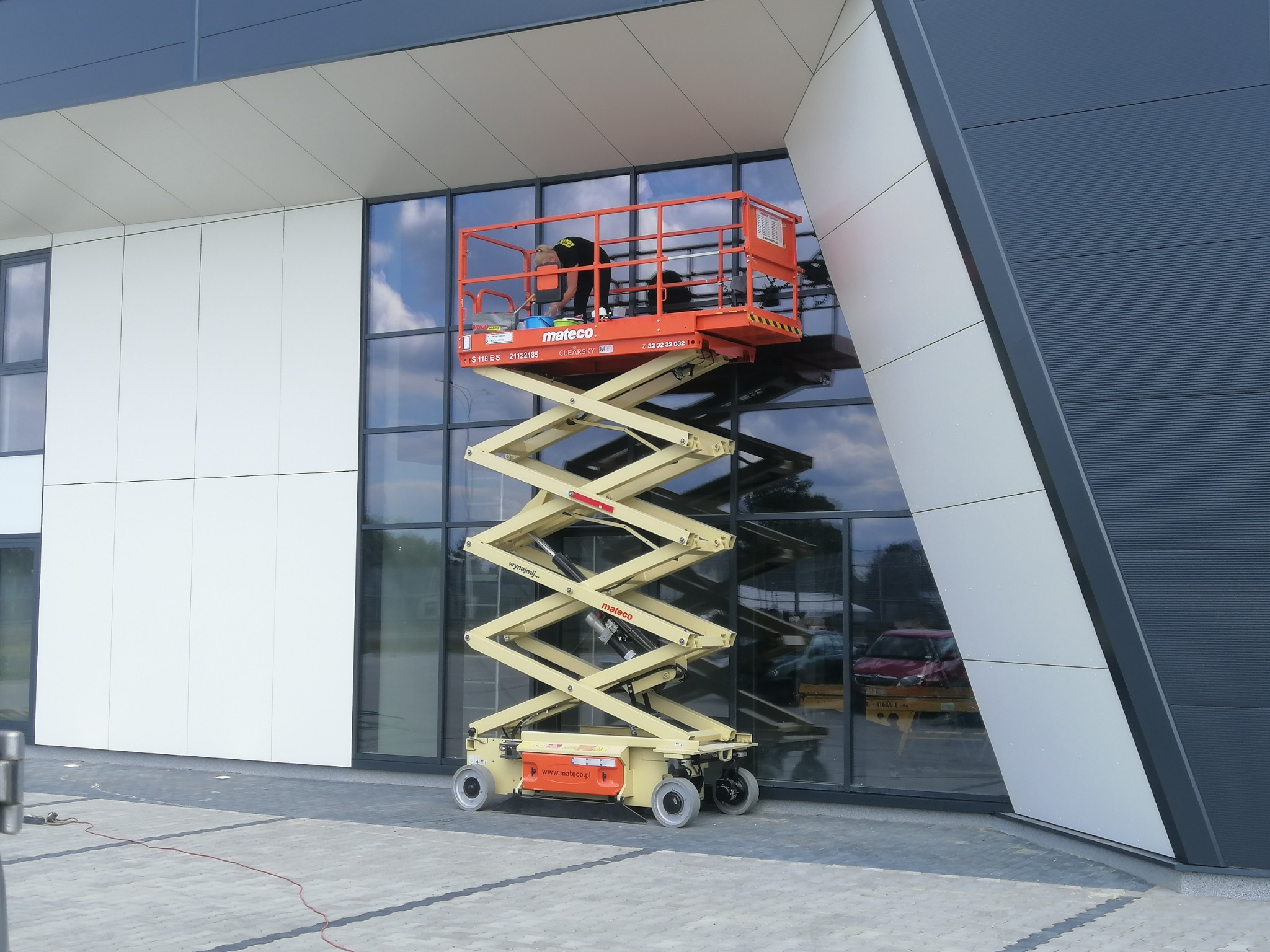 Pracownik w pomarańczowym podnośniku koszowym, myje okna budynku biurowego z szarymi i białymi panelami.