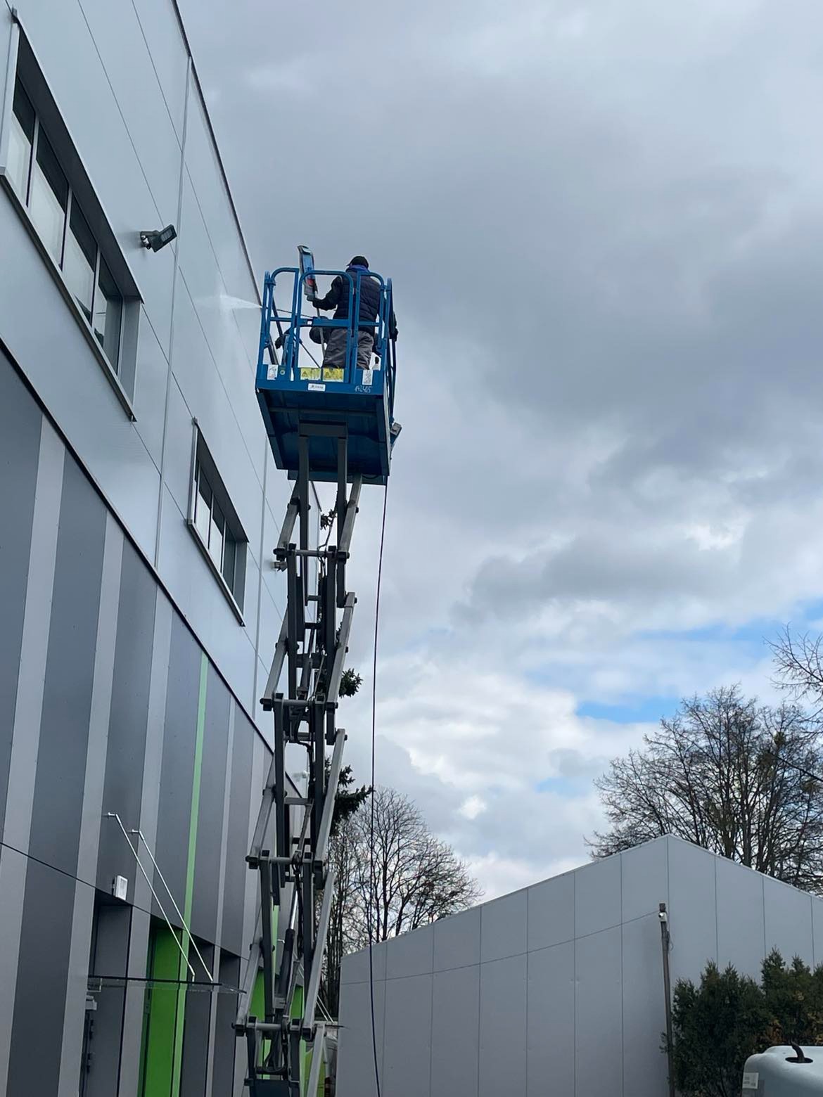 Pracownik na podnośniku koszowym myje elewację budynku biurowego pod ciśnieniem, widoczne okna i lampy na ścianie, pochmurne niebo w tle.