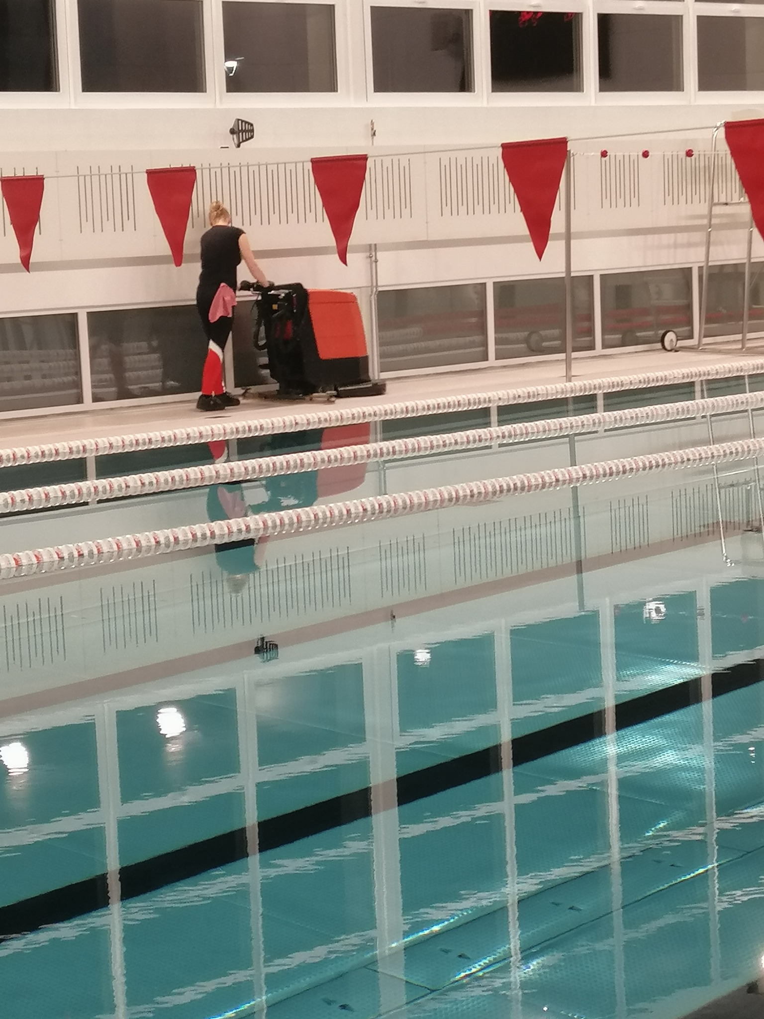 Pracownica w stroju roboczym obsługuje maszynę do czyszczenia podłóg na brzegu basenu, widoczne czerwone flagi i odbicie w wodzie.