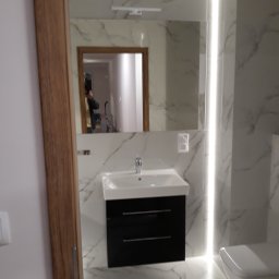 MK RENOV Marek Kondracki - Nowoczesna łazienka z marmurowymi płytkami, podświetlanym lustrem i czarną szafką pod umywalką, widoczne odbicie osoby robiącej zdjęcie w lustrze.