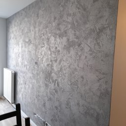 MK RENOV Marek Kondracki - Ściana pokryta dekoracyjną, srebrzysto-szarą farbą o wyraźnej strukturze, widoczna instalacja elektryczna w dolnej części ściany, zabezpieczona podłoga i grzejnik w tle.