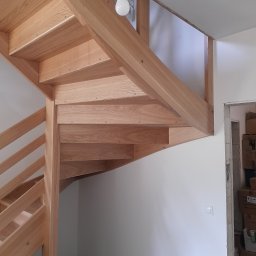 MK RENOV Marek Kondracki - Drewniane schody spiralne z jasnego drewna w trakcie montażu, widoczna surowa żarówka i kartony w tle.