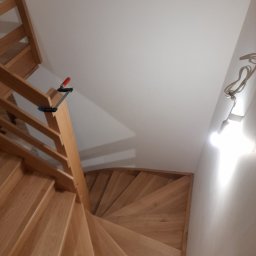 MK RENOV Marek Kondracki - Drewniane schody spiralne z jasnego drewna, widok z góry, z widocznym zaciskiem stolarskim na poręczy i tymczasową żarówką na ścianie, ukazujące etap montażu.