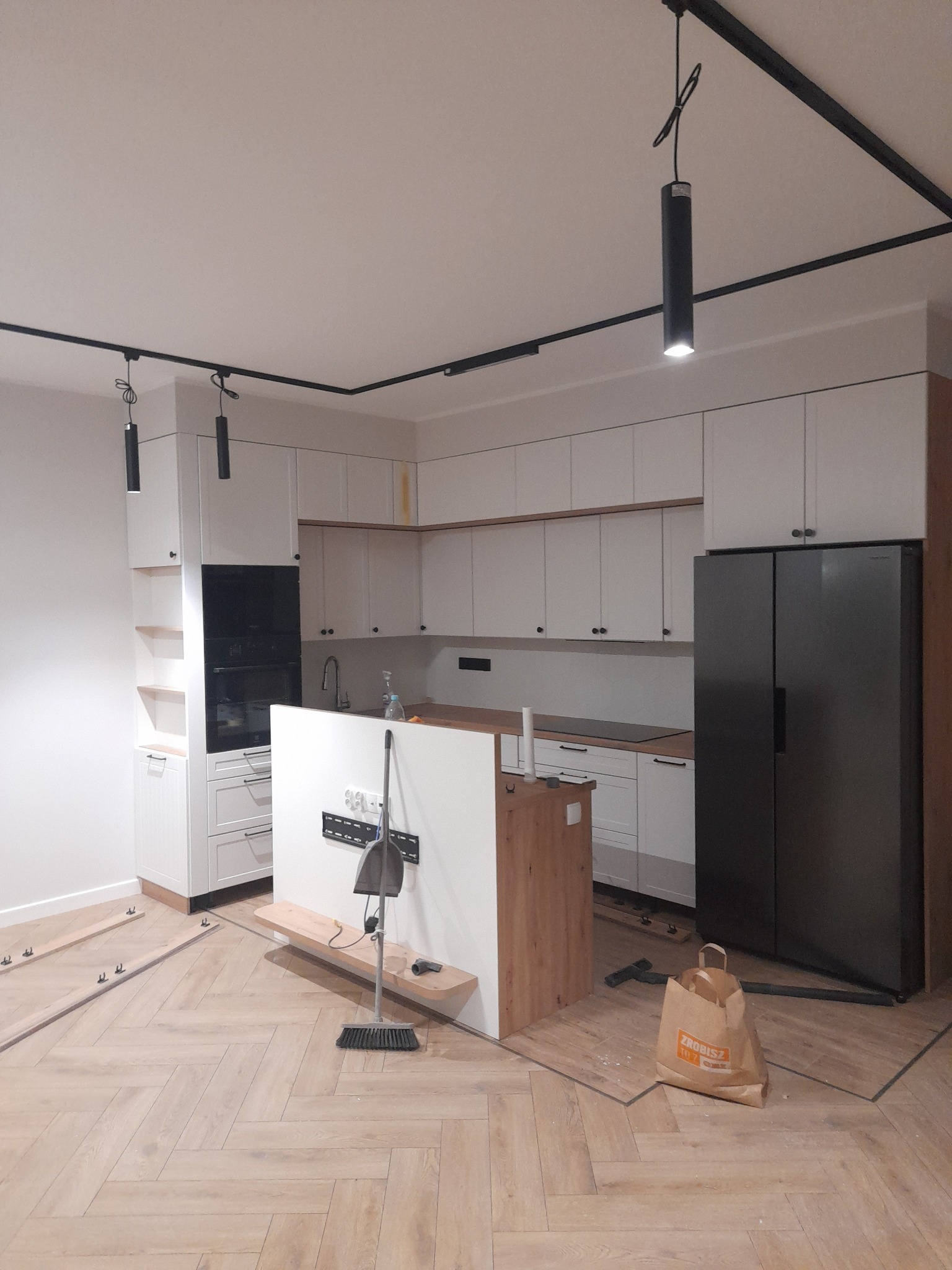 Wnętrze kuchni w trakcie prac wykończeniowych: białe szafki, czarna lodówka, oświetlenie szynowe i parkiet ułożony w jodełkę; widoczne narzędzia i materiały budowlane.