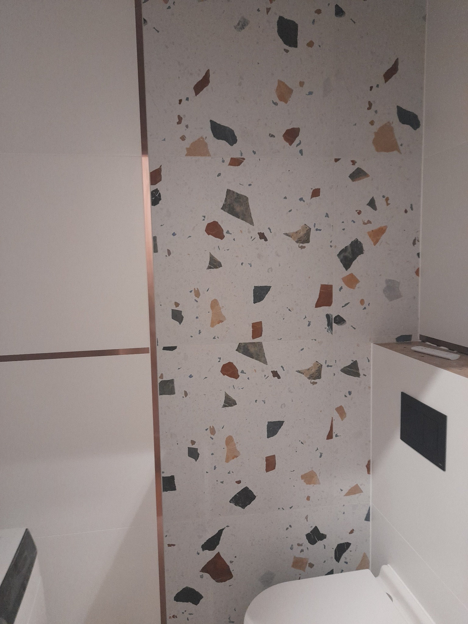 Fragment łazienki z płytkami imitującymi lastryko na ścianie, obok białe panele ścienne z miedzianymi akcentami i zabudowany stelaż podtynkowy toalety.