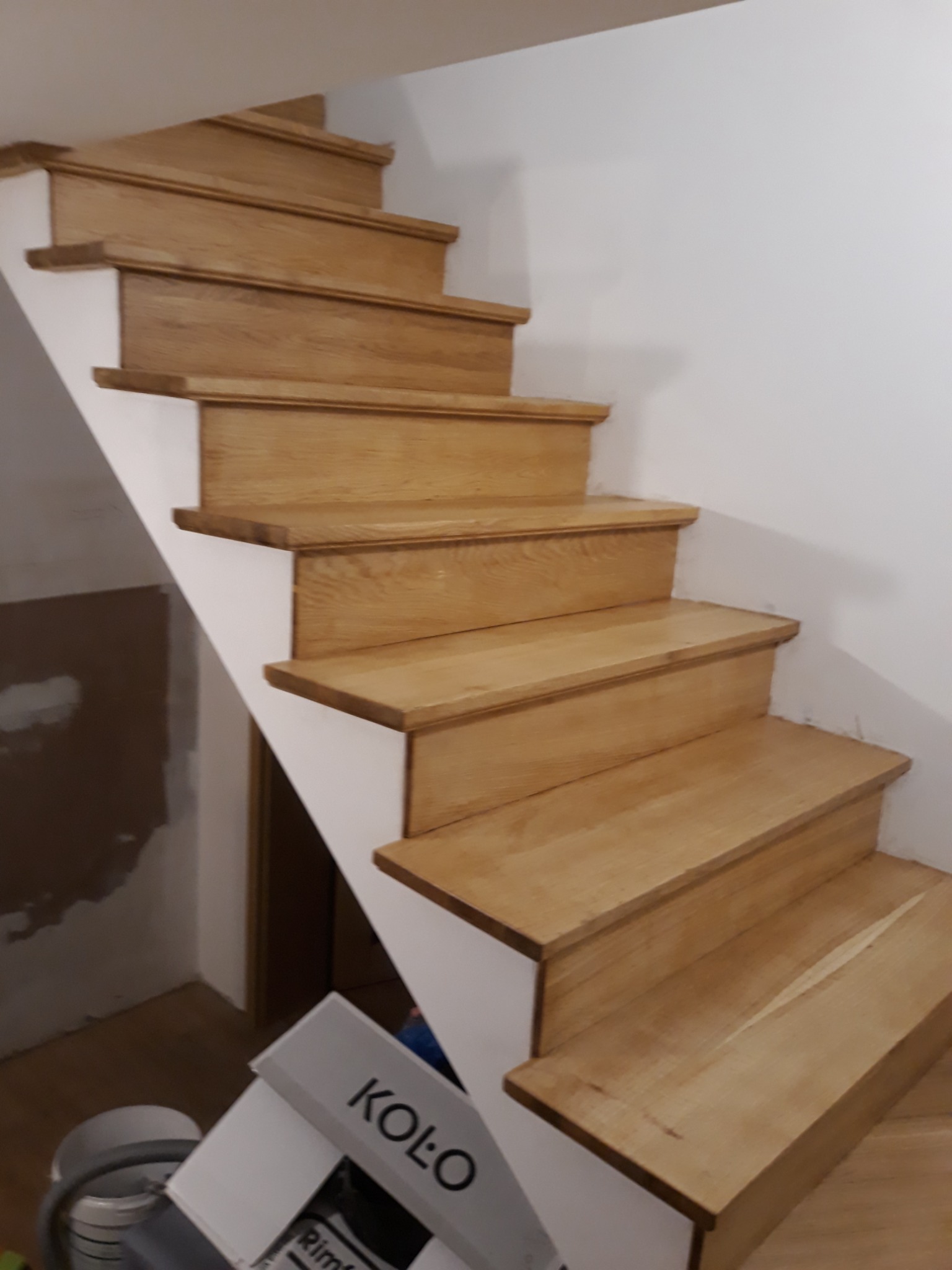 Jasne, drewniane schody z białymi podstopnicami, widoczne z bliskiej perspektywy, z elementami wyposażenia budowlanego w dolnej części kadru.