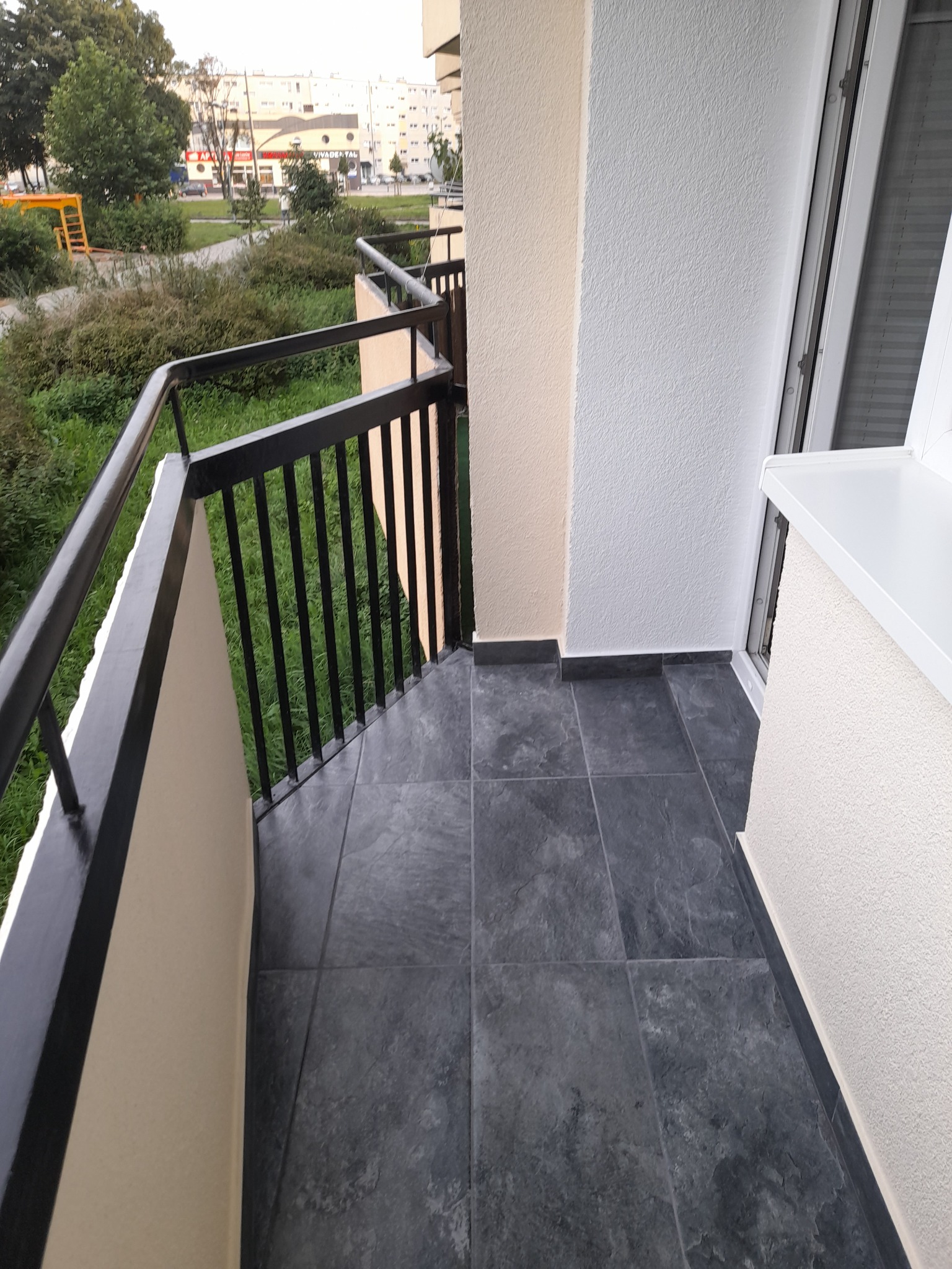 Wykończony balkon z ciemnymi, kwadratowymi płytkami podłogowymi, czarną metalową balustradą i widokiem na zieleń oraz budynki w tle.