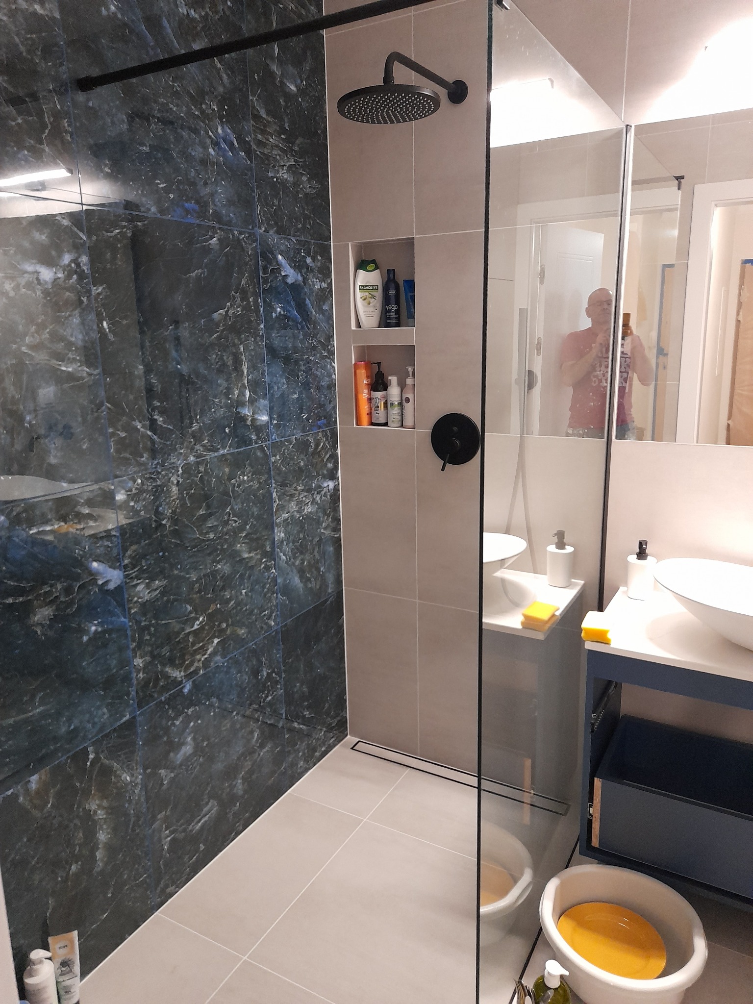 Nowoczesna łazienka po remoncie: kabina typu walk-in z czarną armaturą, ściana wyłożona marmurkowymi płytkami w odcieniach granatu i szarości, wbudowane półki z kosmetykami, umywalki nablatowe...