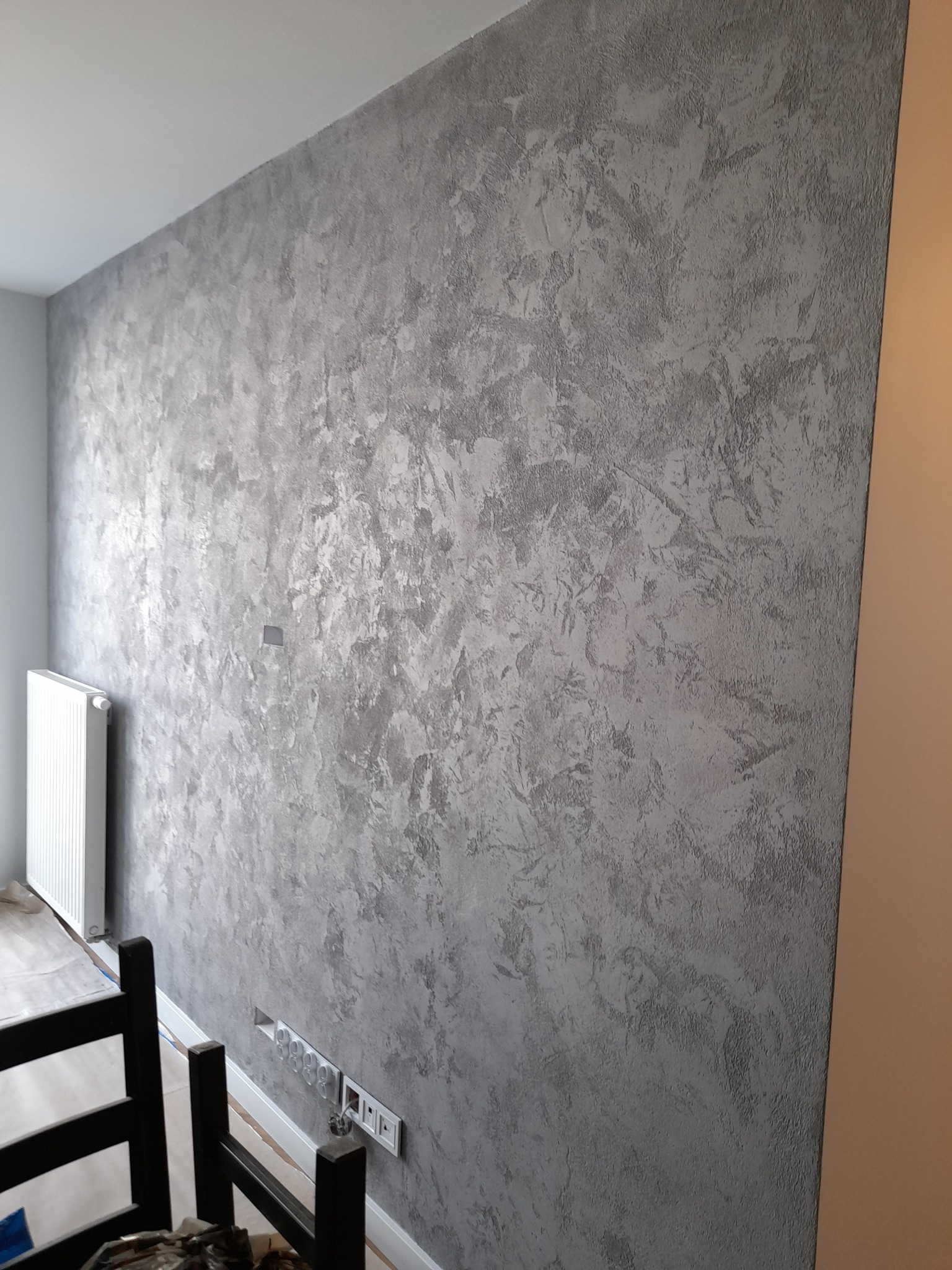 Ściana pokryta dekoracyjną, srebrzysto-szarą farbą o wyraźnej strukturze, widoczna instalacja elektryczna w dolnej części ściany, zabezpieczona podłoga i grzejnik w tle.