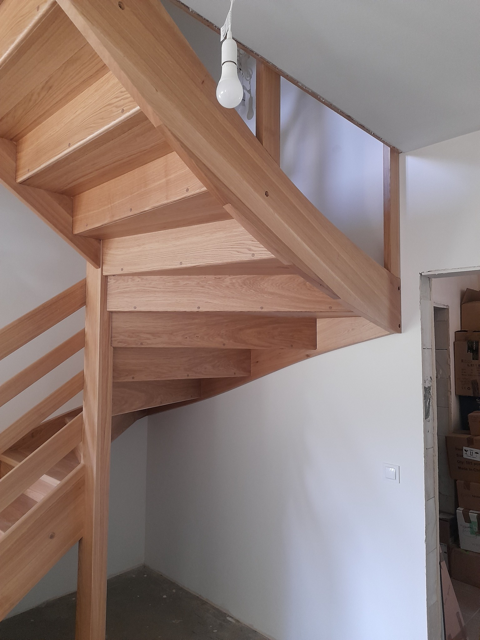 Drewniane schody spiralne z jasnego drewna w trakcie montażu, widoczna surowa żarówka i kartony w tle.