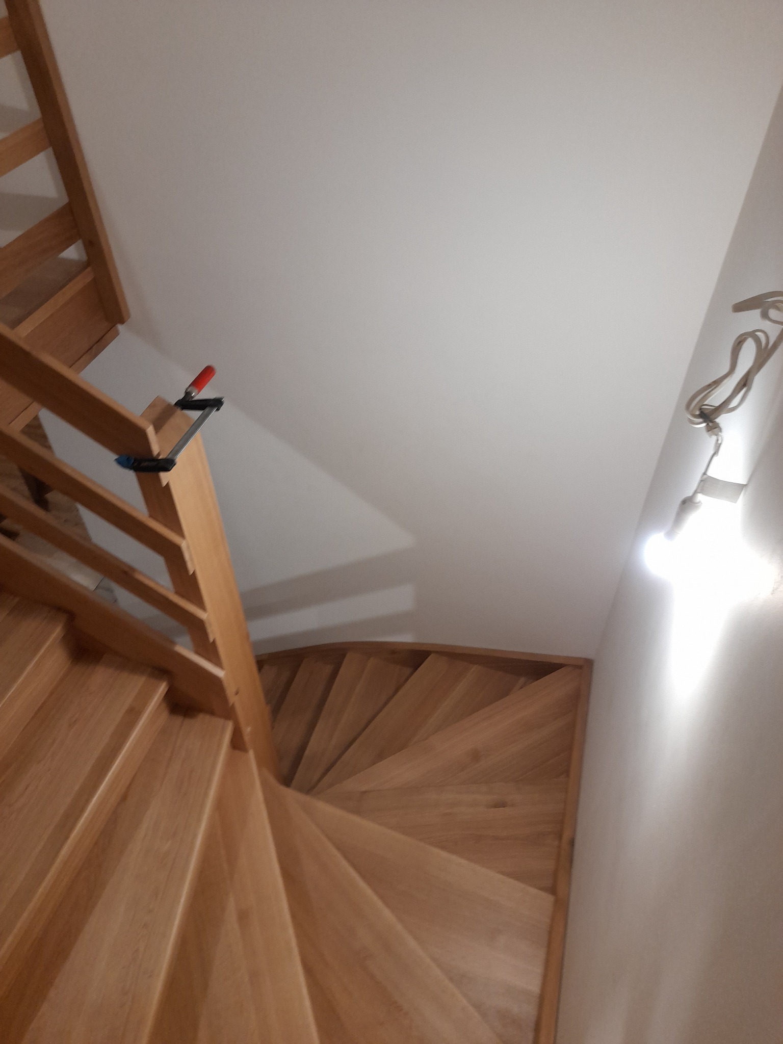 Drewniane schody spiralne z jasnego drewna, widok z góry, z widocznym zaciskiem stolarskim na poręczy i tymczasową żarówką na ścianie, ukazujące etap montażu.