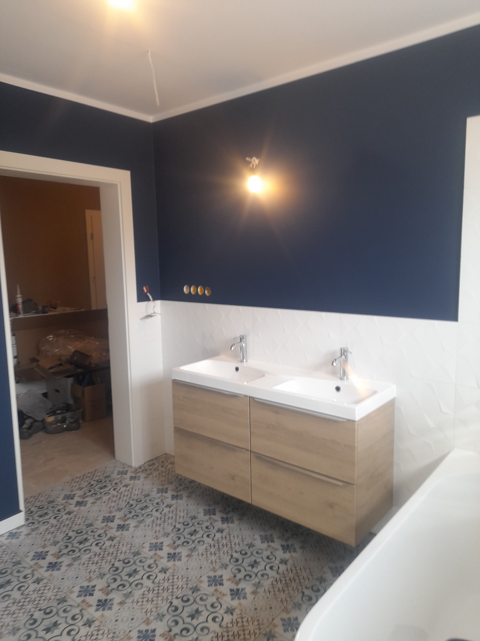 Łazienka w trakcie prac wykończeniowych: podwójna umywalka z szafką w kolorze drewna, wzorzysta podłoga, ściana w kolorze granatowym z białymi płytkami, widoczne przewody elektryczne.