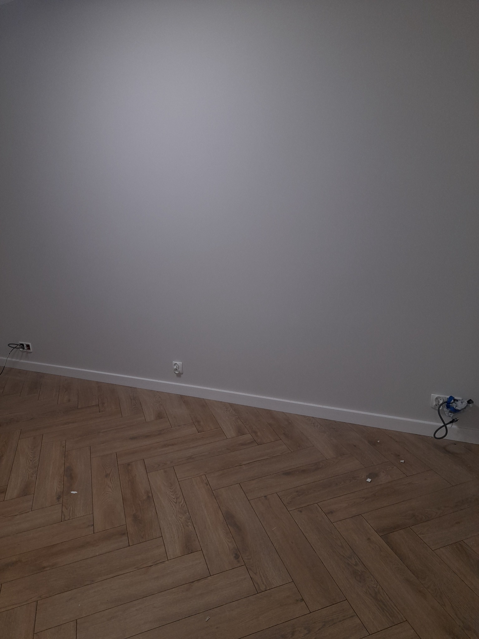 Pokój po remoncie: parkiet ułożony w jodełkę, szara ściana z białymi listwami przypodłogowymi i gniazdkami elektrycznymi.