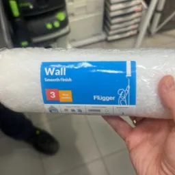 Wałek malarski marki Flügger 'Wall Smooth finish' w folii, trzymany w dłoni na tle sklepowych półek z narzędziami i akcesoriami malarskimi.