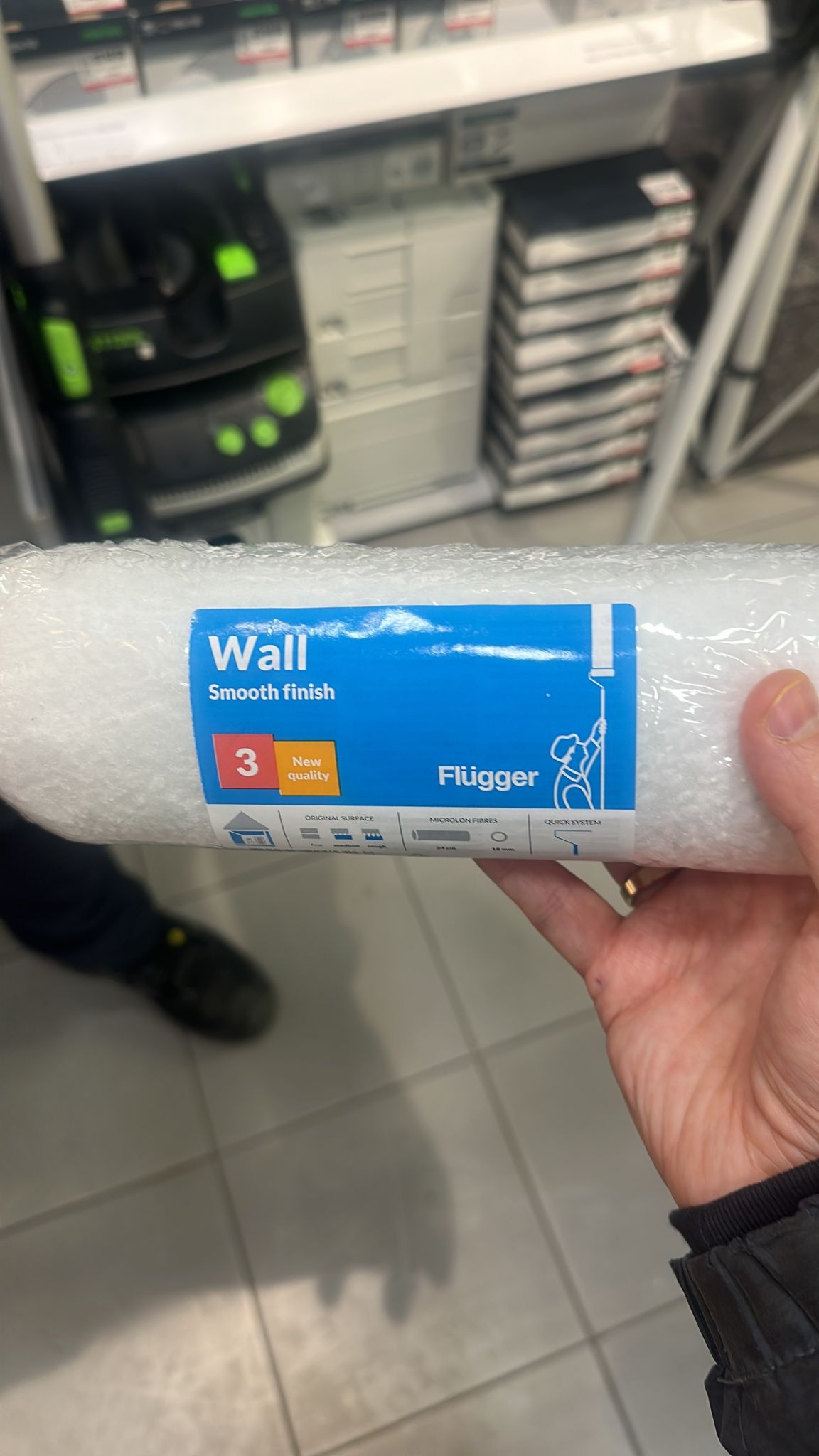 Wałek malarski marki Flügger 'Wall Smooth finish' w folii, trzymany w dłoni na tle sklepowych półek z narzędziami i akcesoriami malarskimi.
