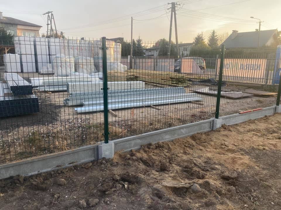 W trakcie instalacji: Fragment ogrodzenia z paneli z betonową podmurówką, widoczne zapasy paneli i słupków w tle.