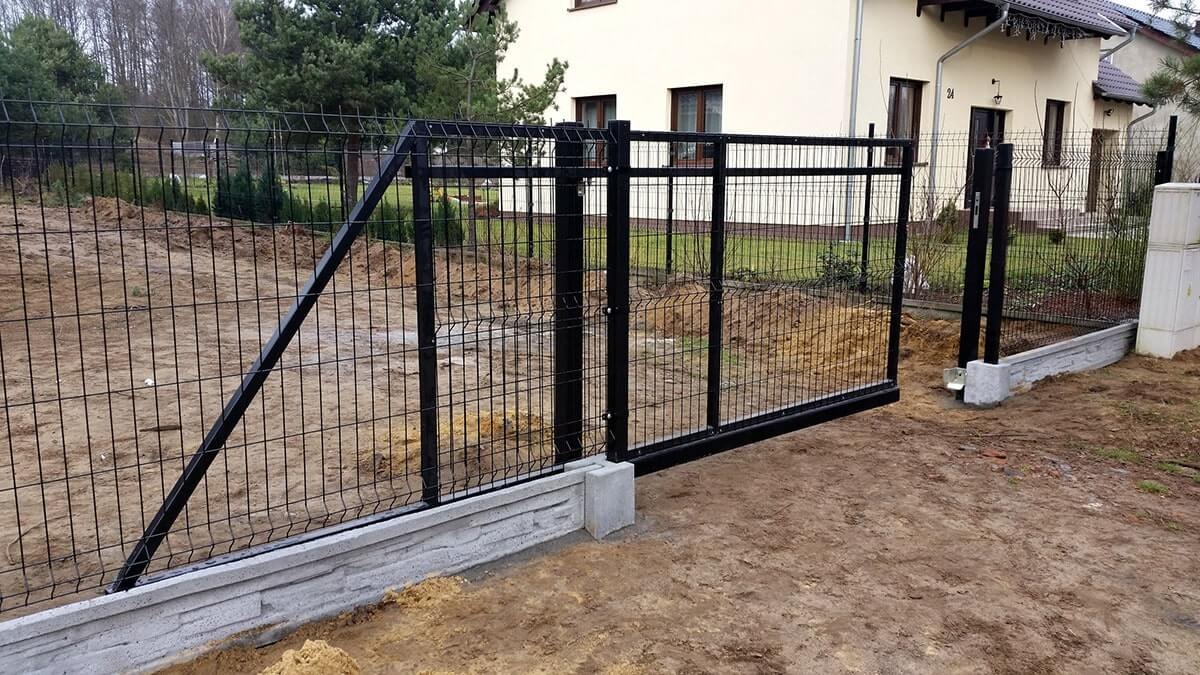 Czarna brama przesuwna i przyległe do niej czarne ogrodzenie z siatki, zamontowane na betonowej podmurówce z elementami dekoracyjnymi imitującymi kamień, na tle budynku mieszkalnego i drzew.