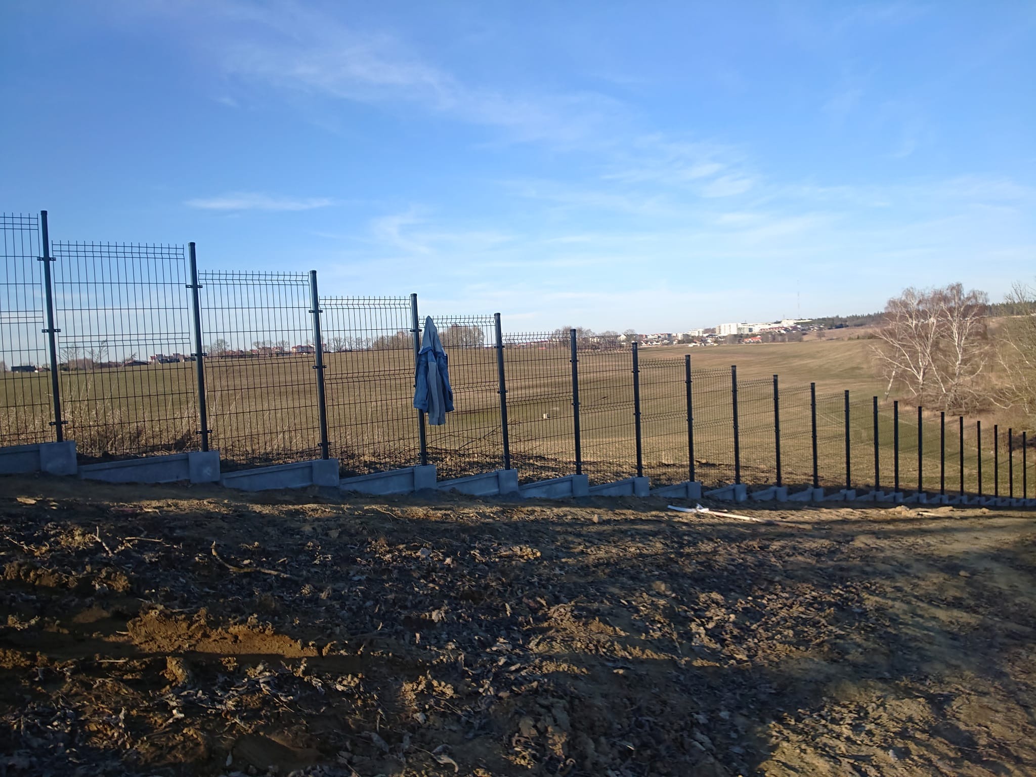 Częściowo zamontowane czarne ogrodzenie z siatki panelowej na betonowej podmurówce, z niebieską kurtką roboczą wiszącą na jednym z paneli, na tle pola i zabudowań miejskich pod błękitnym niebem.