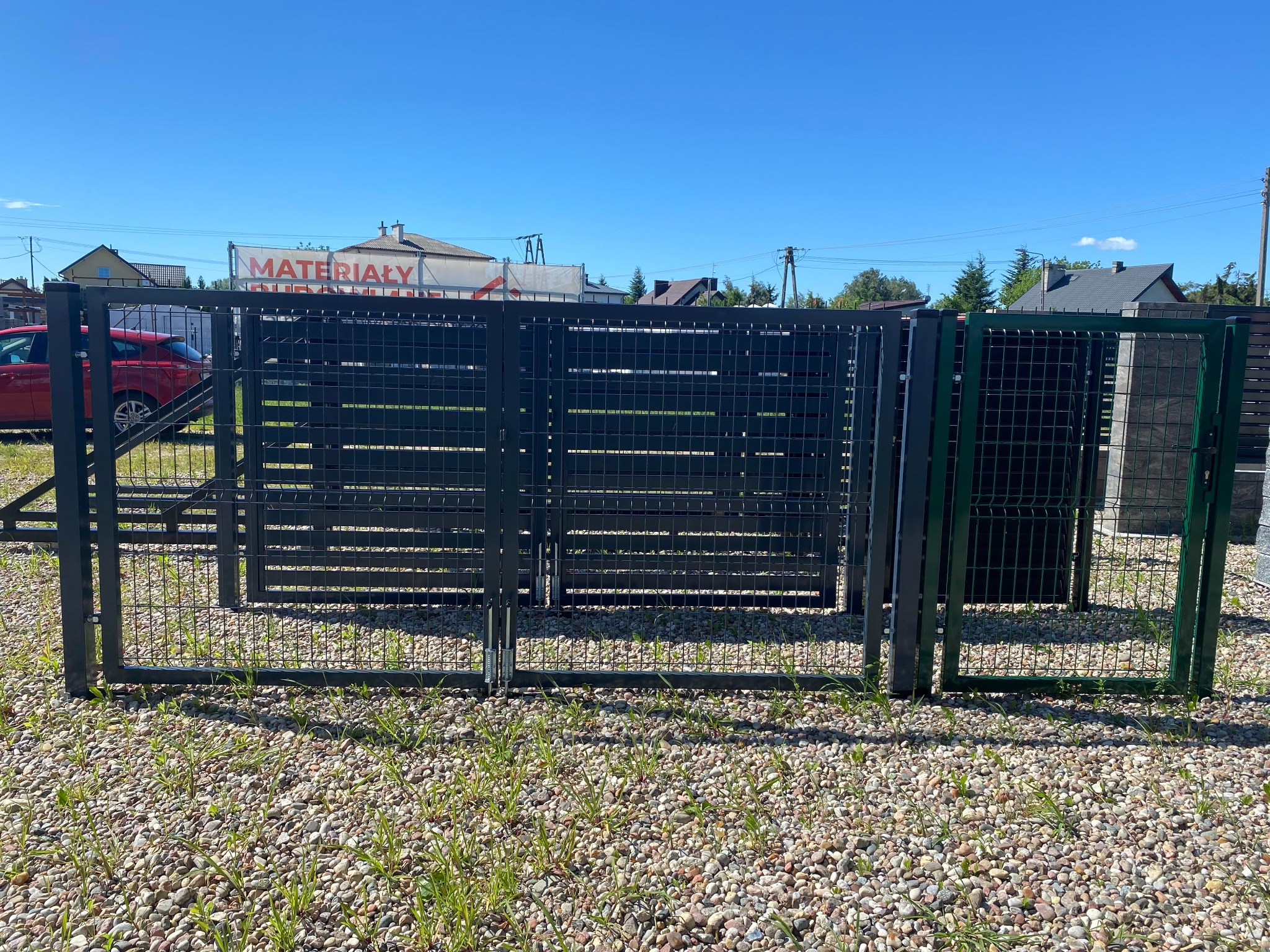 Nowoczesna, czarna, metalowa brama z poziomymi panelami, częściowo ażurowa, ustawiona na żwirowym podłożu z widoczną zielenią, w tle budynki i niebieskie niebo.