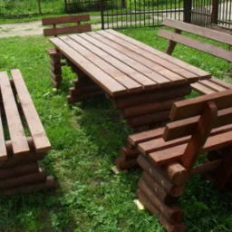 Drewniany stół z ławkami wykonany z okrąglaków, ustawiony na trawie w ogrodzie, widok z boku, ciemne drewno.