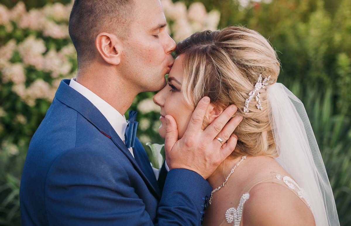 Pocałunek w czoło pary młodej w plenerze, panna młoda z upiętymi blond włosami i ozdobą, pan młody w niebieskim garniturze z muchą, delikatne tło z kwiatów.