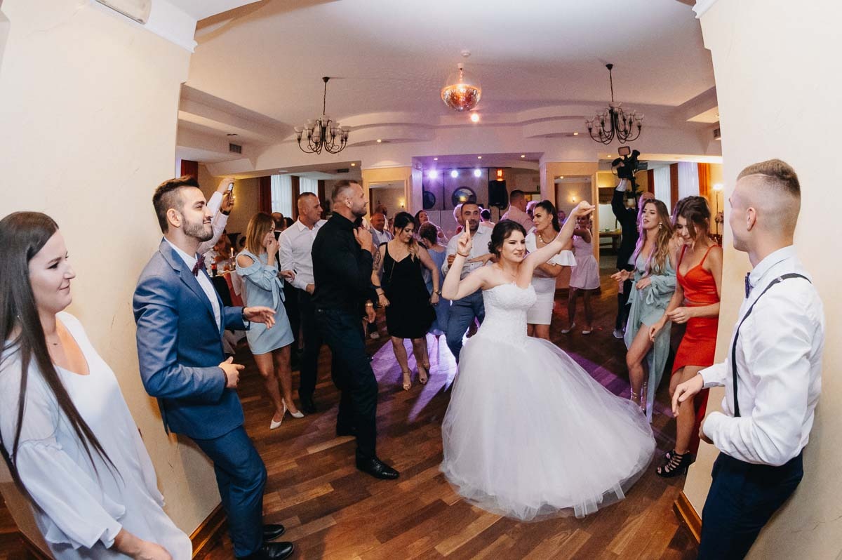 Panna młoda w białej sukni tańczy z gośćmi na parkiecie sali weselnej, oświetlonej żyrandolami i dyskotekową kulą.