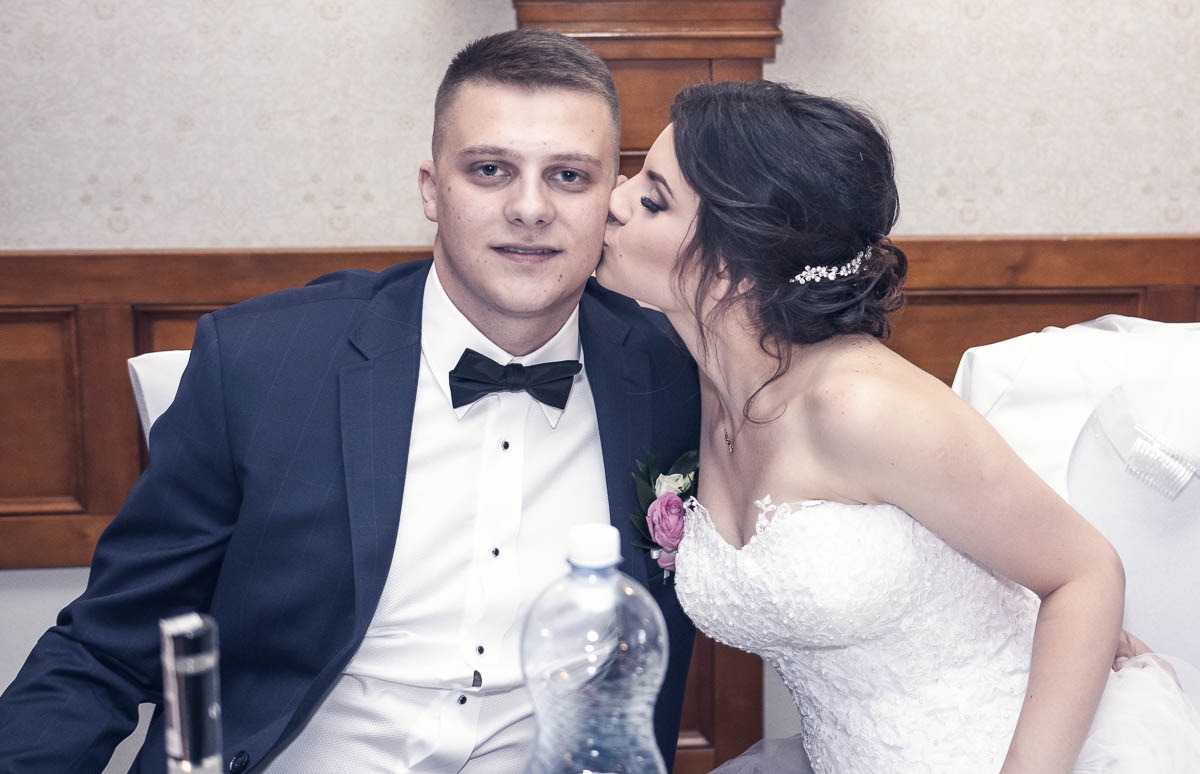 Panna młoda w koronkowej sukni całuje pana młodego w ciemnym garniturze z muchą, widoczna butelka wody na stole.