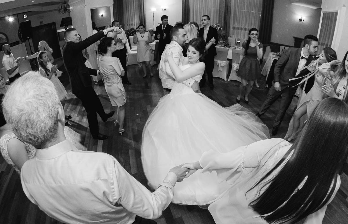Czarno-białe zdjęcie pary młodej tańczącej w otoczeniu gości weselnych na parkiecie z drewnianych desek, z widocznymi stołami i krzesłami w tle.