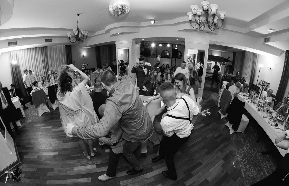 Czarno-białe zdjęcie z szerokim kątem przedstawiające tańczących gości na parkiecie podczas uroczystości, z widocznymi nakrytymi stołami i dekoracjami w tle.