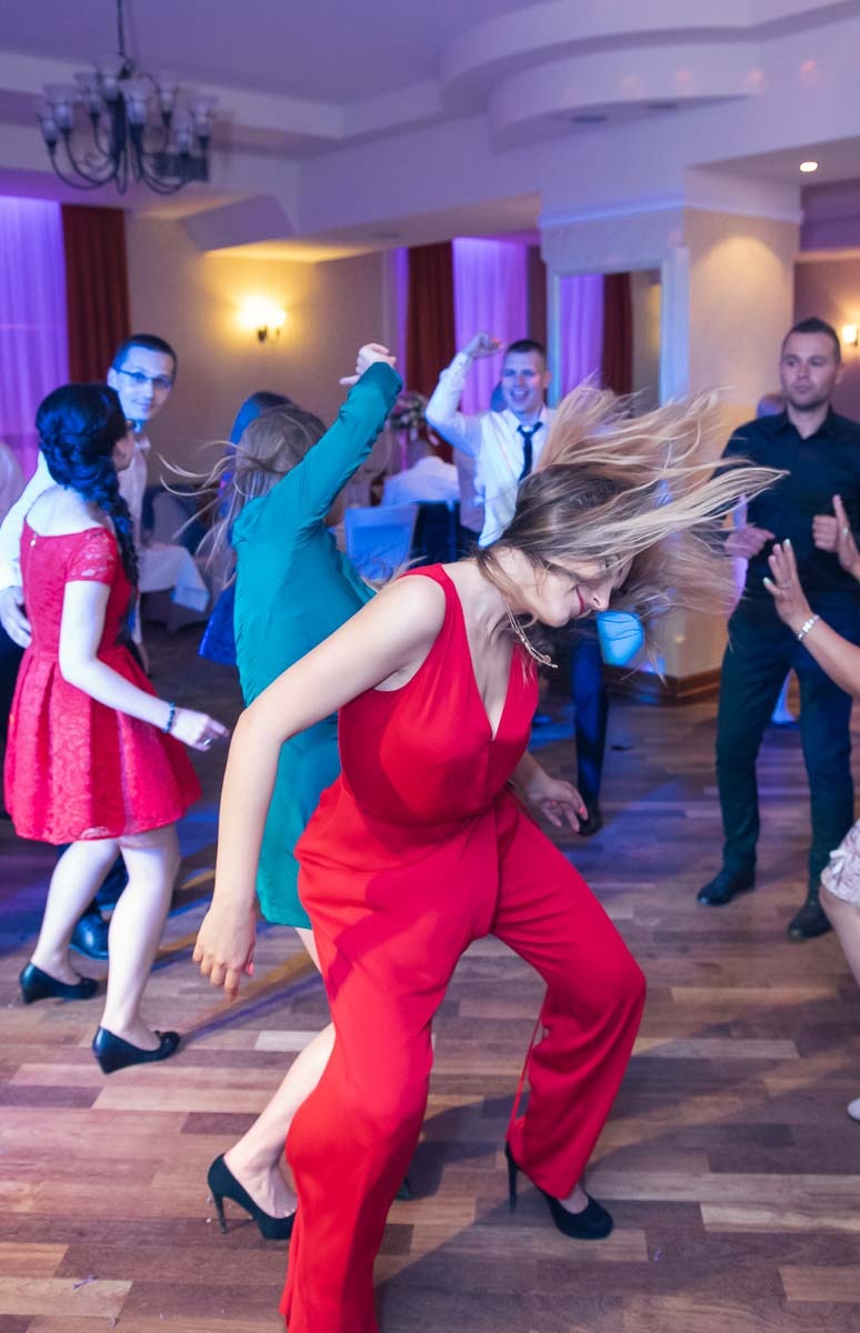 Roztańczona kobieta w czerwonym kombinezonie z szerokimi nogawkami, włosy w ruchu, w tle tańczący goście w sali z parkietem i fioletowym oświetleniem.