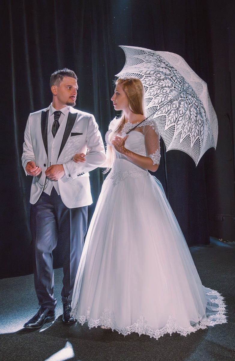 Elegancka para: mężczyzna w białym smokingu z czarną kamizelką i kobieta w białej sukni ślubnej z koronkową parasolką na tle czarnych zasłon.