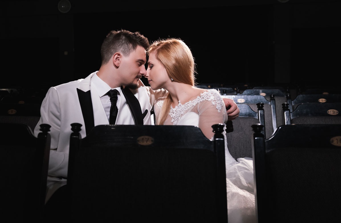 Para młoda w kinie: Panna młoda w sukni z koronkowymi rękawami i pan młody w białym smokingu, siedzą w ciemnych fotelach kinowych, zbliżając się do siebie twarzami.