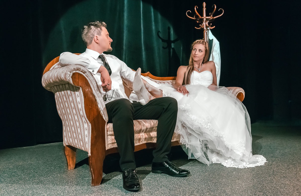 Para młoda, on z zegarkiem kieszonkowym, siedząca na stylizowanej kanapie na tle ciemnej sceny z wieszakiem z marynarką.